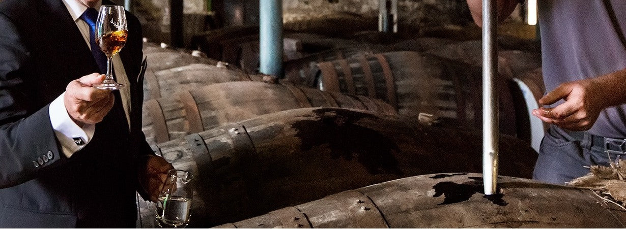 Taça Copita com whisky The Dalmore sendo avaliada no armazém, ao lado de barrica sendo aberta